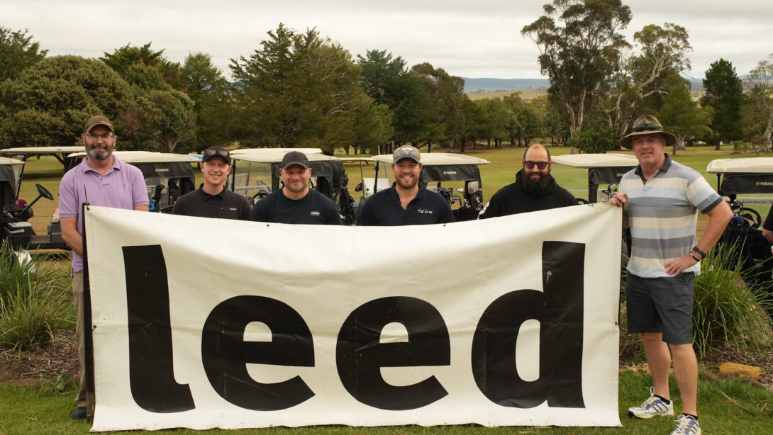 Leed - A group of Leed Engineering staff holding a large Leed banner at a charity golf day supporting the community in Cooma.