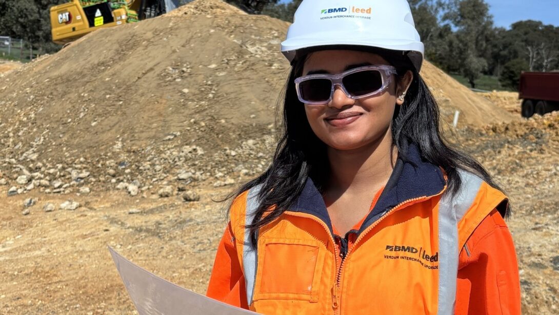 Leed - Leed Safety Advisor Monicka Parameshwaran is pictured in a high vis jacket and hard hat. She is holding a page and is in front of a mount of dirt and an excavator on the Verdun Interchange site